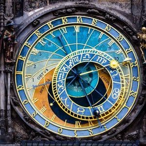 Un primer plano de un reloj astronómico ornamentado con esferas azules y doradas, signos del zodiaco y figuras talladas en un edificio de piedra.