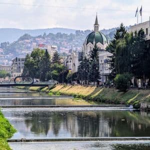 Un fiume scorre su piccoli sbarramenti attraverso una città con un edificio dalla cupola verde e una collina ricoperta di case.