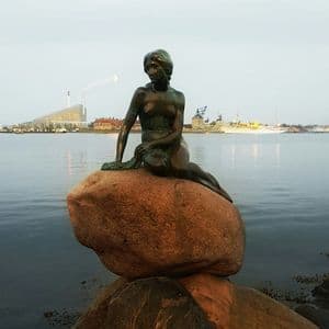 Una statua di bronzo di una sirena seduta su una grande roccia in riva all'acqua, con uno skyline cittadino visibile sullo sfondo al tramonto.
