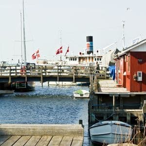 Segelboote mit dänischen Flaggen liegen in einem sonnigen Hafen neben einem roten Holzgebäude und einer Fußgängerbrücke.