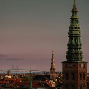 Veduta della città al tramonto con ornate torri di chiesa e un ponte sospeso in lontananza.
