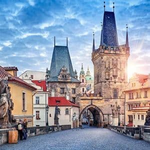 Vista desde un puente histórico de adoquines con estatuas, hacia una torre medieval al amanecer bajo un cielo nublado.