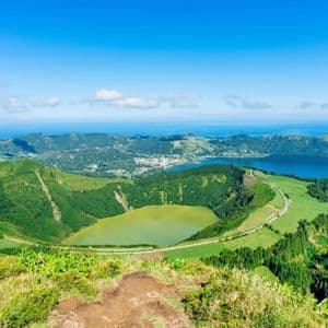 Vue plongeante sur des collines volcaniques vertes entourant deux lacs de cratère, l'un vert et l'autre bleu, avec l'océan à l'horizon.