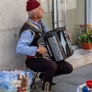 Un uomo con un fez rosso e un gilet tradizionale suona una fisarmonica nera mentre è seduto su uno sgabello su un marciapiede.
