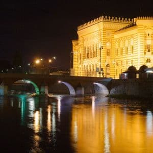 Un sontuoso edificio e un ponte ad arco in pietra sono illuminati di notte, le loro luci si riflettono su un ampio fiume.