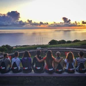 Un grupo de WeRoad se sienta en un muro de piedra, observando el atardecer sobre el océano desde un mirador verde.