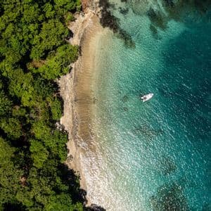 Una vista aérea de un barco blanco flotando en agua clara y turquesa junto a una playa de arena bordeada por un denso bosque verde.
