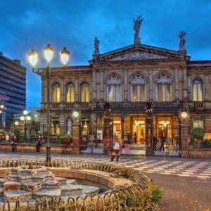 Le bâtiment orné du Théâtre National du Costa Rica, illuminé au crépuscule, avec une fontaine décorative au premier plan d'une place de ville.
