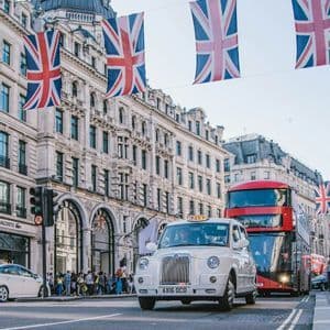 Un taxi londonien blanc et un bus à impériale rouge circulent dans une rue décorée de drapeaux Union Jack suspendus entre des bâtiments ornés.