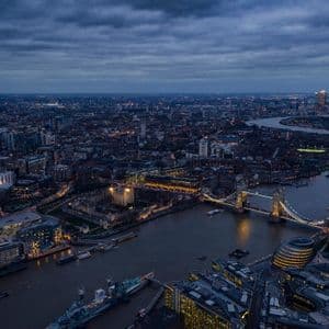 Una vista aerea del panorama urbano di Londra al tramonto, con il Tower Bridge illuminato e il Tamigi sotto un cielo nuvoloso.
