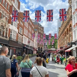 Una strada cittadina affollata con persone che camminano e cenano all'aperto, decorata con festoni di bandiere Union Jack appesi tra edifici in mattoni.