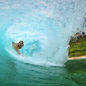 Un surfista cavalca all'interno del tubo di una grande onda turchese, con la vista che si apre verso una spiaggia sabbiosa e una costa verde.