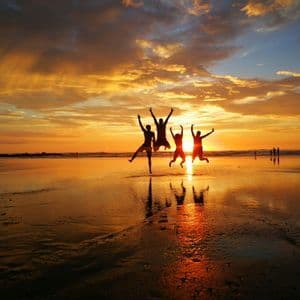 Sagome di quattro persone di un viaggio di gruppo WeRoad che saltano su una spiaggia al tramonto dorato, con riflessi sulla sabbia bagnata.