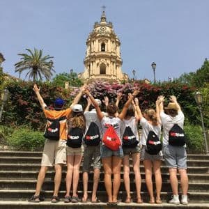 Un voyage de groupe WeRoad vu de dos, debout sur des marches en pierre, les bras levés vers une église ornée d'un clocher.