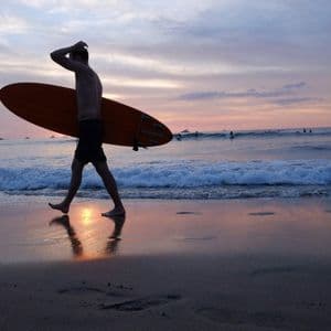 Una silhouette di un uomo con la tavola da surf cammina lungo la riva bagnata di una spiaggia durante un tramonto colorato.