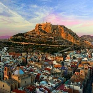 Veduta aerea di una città costiera con una fortezza in cima a una collina, sotto un cielo al tramonto dai colori vivaci.