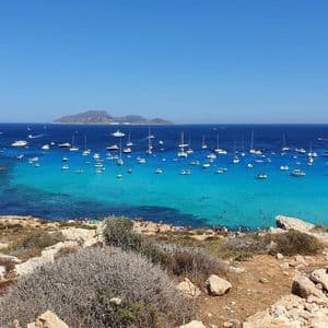 Vista dall'alto di una costa rocciosa con baia turchese e numerose barche ancorate, e un'isola in lontananza.