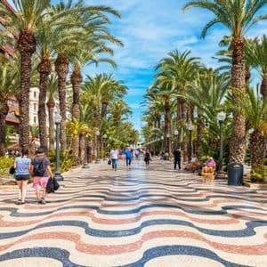 Le persone passeggiano lungo un ampio lungomare fiancheggiato da alte palme, caratterizzato da una pavimentazione a mosaico colorata e ondulata sotto un cielo azzurro.