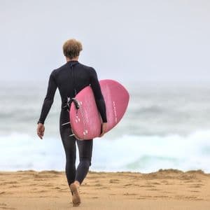 Da dietro, un surfista in muta nera porta una tavola da surf rosa mentre cammina su una spiaggia sabbiosa verso l'oceano.