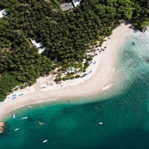 Una vista aerea di una spiaggia di sabbia bianca, delimitata da una lussureggiante foresta verde e acque turchesi cristalline, con piccole barche che galleggiano nelle vicinanze.