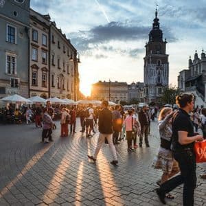 Eine Menschenmenge flaniert bei Sonnenuntergang über einen historischen europäischen Kopfsteinpflasterplatz, mit einem markanten Glockenturm im Hintergrund.