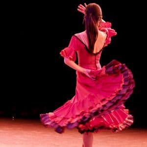 Una ballerina di flamenco, ripresa da dietro, si esibisce su un palco, il suo abito rosa e viola a balze che turbina con il movimento.