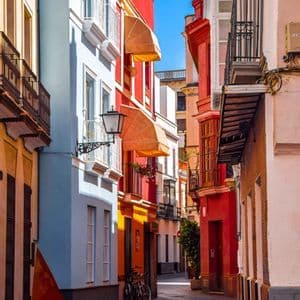 Un vicolo stretto e lastricato è fiancheggiato da vivaci edifici rossi, blu e gialli con piccoli balconi sotto un cielo sereno.