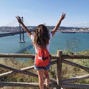 Une femme vue de dos, portant un sac à dos WeRoad rouge, lève les bras en surplombant un grand pont et une ville au-delà de l'eau.