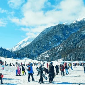 Numerosi sciatori e snowboarder su una pista affollata ai piedi di montagne innevate sotto un cielo blu.
