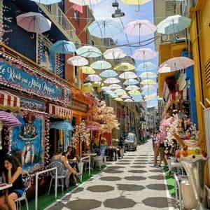 Sombrillas de colores pastel cuelgan suspendidas sobre una calle estrecha con cafés al aire libre, proyectando sombras distintas sobre el pavimento.