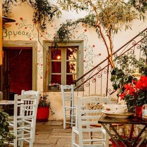 Sillas blancas dispuestas en el patio de piedra de un café griego tradicional, con diseños florales pintados y enredaderas en las paredes.