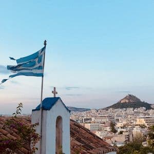 Una bandera griega ondea al viento sobre un campanario blanco con techo azul, con vistas a un denso paisaje urbano y una gran colina al anochecer.