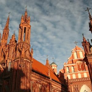 Una vista dal basso di un'ornata chiesa gotica in mattoni rossi, con molteplici guglie, stagliata contro un cielo blu con nuvole bianche.