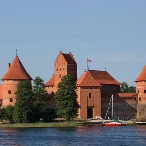 Un grande castello in mattoni rossi con torri coniche si erge sulla riva di un lago, con un pedalò sull'acqua in primo piano.