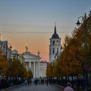 Un'ampia strada cittadina costeggiata da alberi autunnali porta a una grande cattedrale e campanile al crepuscolo, con persone che passeggiano lungo il marciapiede.