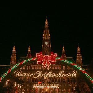 L'arco d'ingresso del Wiener Christkindlmarkt, adornato con un fiocco rosso, risplende di notte davanti a un edificio sontuoso, con una folla sottostante.