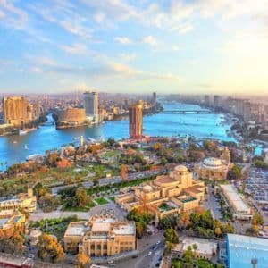 Una vista aerea di una vasta città tentacolare attraversata da un ampio fiume blu, fiancheggiata da edifici moderni e parchi verdi sotto un cielo soleggiato.