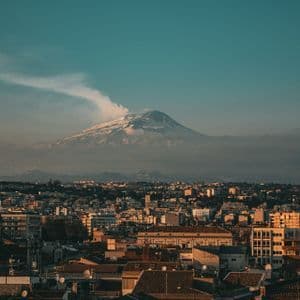 Un paesaggio urbano sotto un vulcano innevato, con un pennacchio di fumo contro un cielo color ottanio.