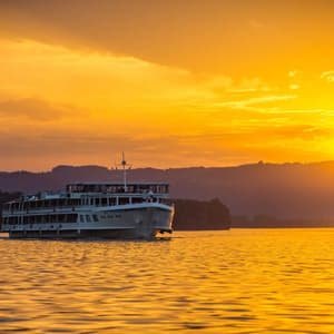 Un crucero fluvial blanco navega sobre aguas tranquilas durante una vibrante puesta de sol dorada, con el sol reflejándose en la superficie.