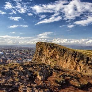A panoramic view from a high, rocky cliff overlooking a sprawling city and a distant body of water under a blue sky with clouds.