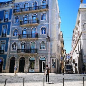 Un alto edificio esquinero con una fachada de azulejos azules y blancos estampados se alza en una calle empedrada bajo un cielo azul claro.