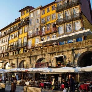 Edificios coloridos y con azulejos se alzan sobre una arcada de piedra con cafés al aire libre en una plaza de la ciudad, bajo un cielo azul claro.