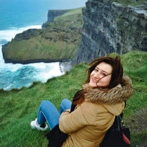 Une femme souriante en manteau d'hiver est assise au bord d'une falaise herbeuse, surplombant une mer agitée et des formations rocheuses côtières spectaculaires.