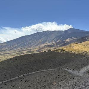 Un viaggio di gruppo WeRoad fa trekking su un sentiero che attraversa un oscuro paesaggio vulcanico, con un vulcano fumante sullo sfondo sotto un cielo azzurro.