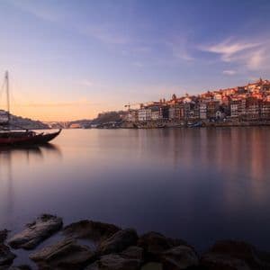 Vista de larga exposición de un barco tradicional en un río tranquilo, frente a una ciudad histórica en una colina bajo un colorido cielo al atardecer.