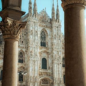The ornate white facade of a Gothic cathedral with numerous spires, framed between two large, carved stone columns.