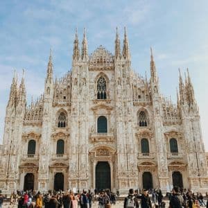 The ornate, spired facade of a large gothic cathedral with a crowd of people and pigeons gathered in the square below.