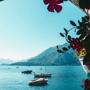 A view of a large lake with several boats, seen from under a stone archway with pink flowers in the foreground and mountains in the background.