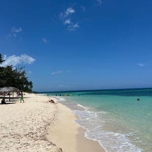 Una playa de arena blanca se encuentra con aguas cristalinas de color turquesa bajo un cielo azul brillante, con algunas personas nadando y relajándose.