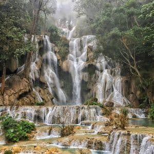 Una cascata a più livelli scende lungo sporgenze rocciose in pozze turchesi, circondata da una foresta verde e nebbiosa.